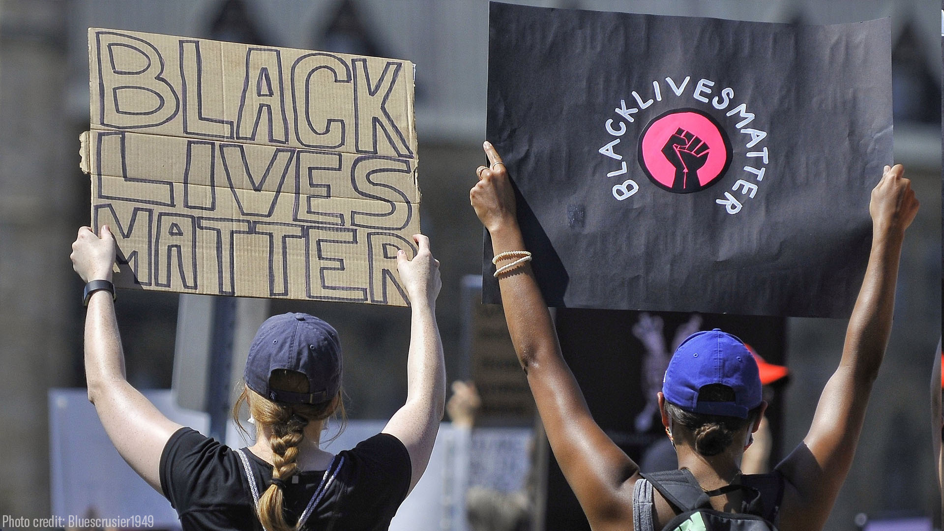 Photo: Two people are pictured with their back facing the camera. They are outside rand raise handwritten signs that say "Black Lives Matter."