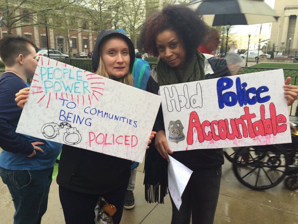 Two women are at a rally. The woman on the right has a sign that says, "People Power to Communities being policed" and the woman one the left has a sign that says, "Hold police accountable."