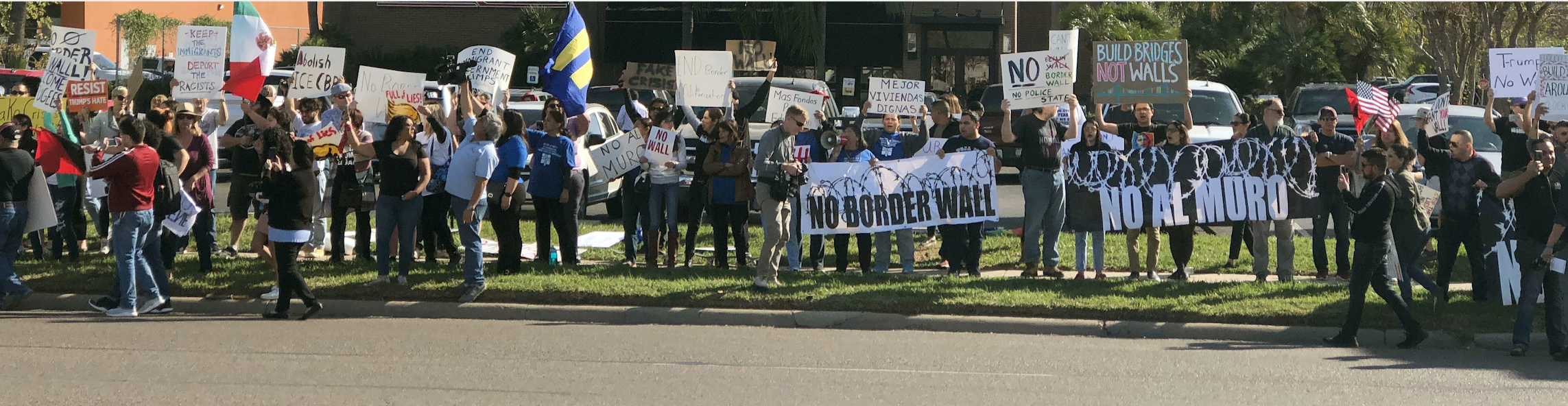 Photo: A crowd of people stand along a street in the Rio Grande Valley. Behind them is a restaurant and other businesses. They hold signs, one of which says "No Border Wall."